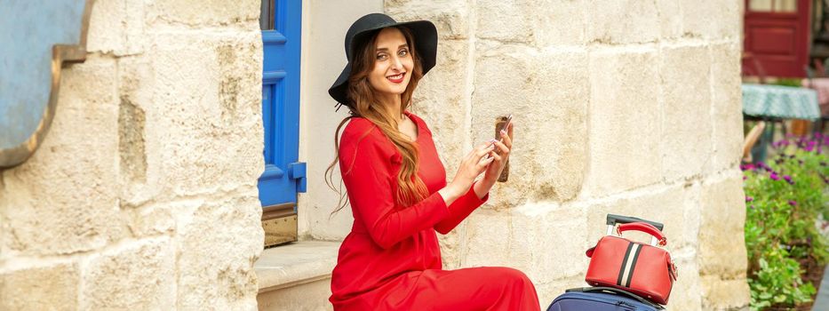 Portrait of tourist young caucasian woman in a red long dress and black hat with suitcase sitting on the stairs outdoors