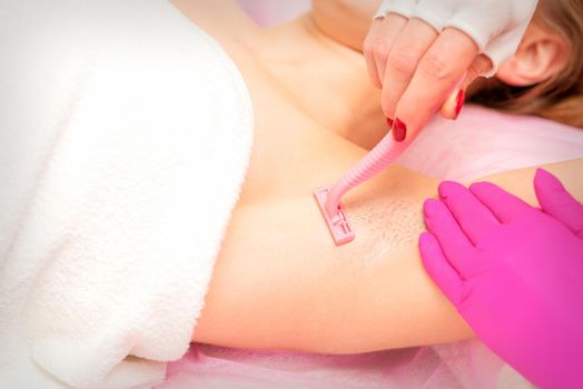 Beautician shaves the armpit of a young woman with a razor before the hair removal procedure