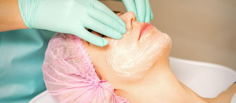 A cosmetologist is applying cream on the female face, close-up view. Woman with doctor beautician in beauty clinic