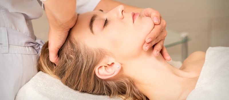 Cosmetologist hands doing facial massage on forehead and chin of young female at spa salon