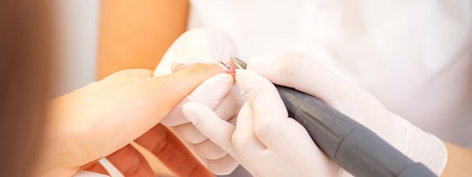 Closeup of manicure master in white gloves applying an electric nail file drill to remove the nail polish in the beauty salon