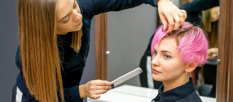 Female hairdresser styling short pink hair of the young white woman with hands and comb in a hair salon