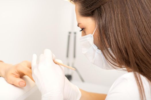 Manicure master in protective face mask and white gloves apply polish to female fingernails