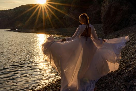 A mysterious female silhouette with long braids stands on the sea beach with mountain views, Sunset rays shine on a woman. Throws up a long white dress, a divine sunset