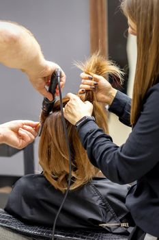 The rear view of two hairdressers are curling hair for a young woman with electric hair iron in a beauty salon