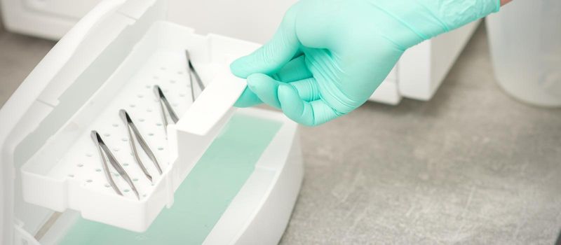 The beautician holds a tray of tweezers preparing them for disinfection with a special liquid and machine