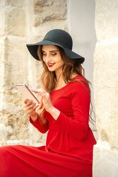 A young traveling white woman in the red jacket and black hat sitting with a smartphone on the city street