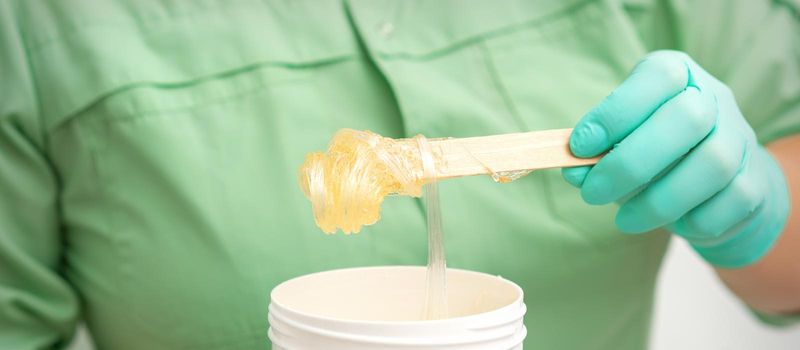 Liquid yellow sugar paste for depilation on a stick flows into the jar in hands of a beautician