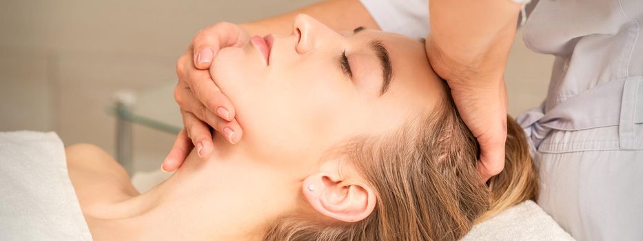 Cosmetologist hands doing facial massage on forehead and chin of young female at spa salon