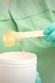 Liquid yellow sugar paste for depilation on a stick flows into the jar in hands of a beautician