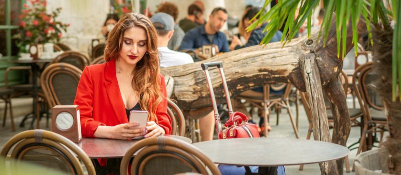 Tourist young caucasian white woman in a red jacket looking at smartphone sitting at the table in cafe outdoors