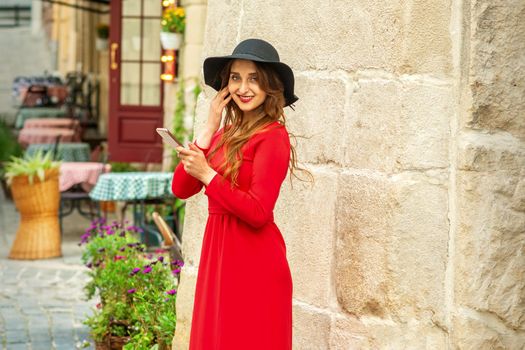 A young traveling white woman in the red jacket and black hat standing with a smartphone on the city street