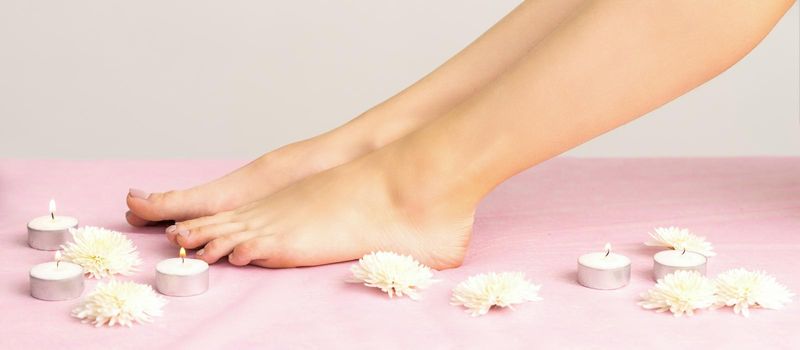 Beautiful white smooth feet of young woman with candles and flowers on pink couch