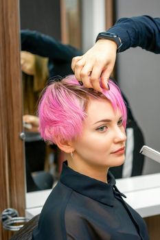 Female hairdresser styling short pink hair of the young white woman with hands and comb in a hair salon