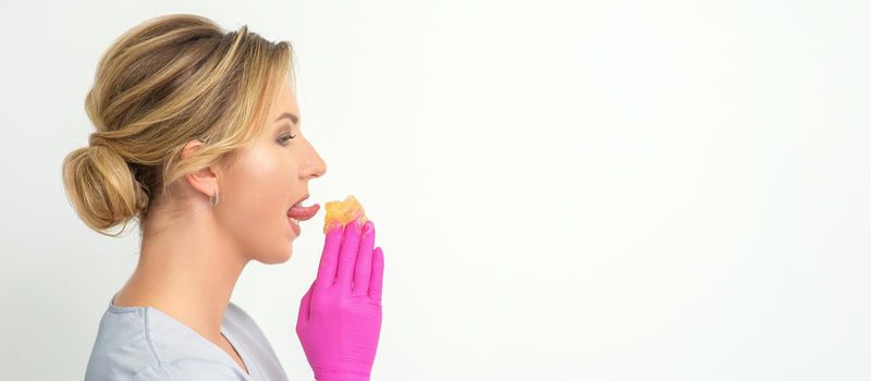 Young beautician licks liquid wax for depilation holding in gloved hands standing on white background. Cosmetologist tastes the sugar paste to taste