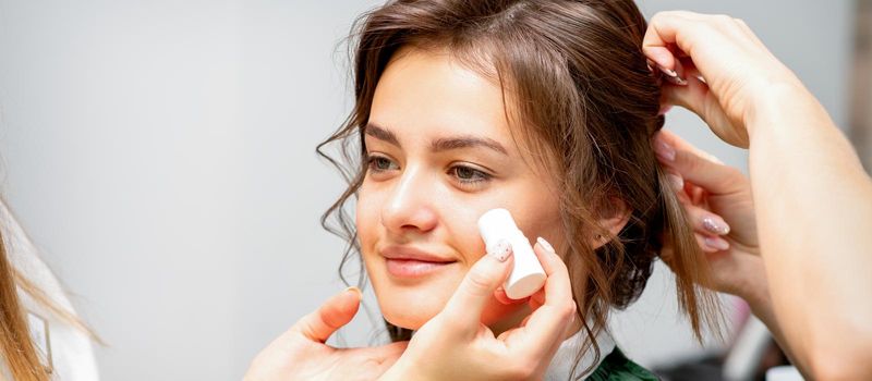 Makeup artist applying cream blush foundation tube on the cheek of the young caucasian woman in a beauty salon