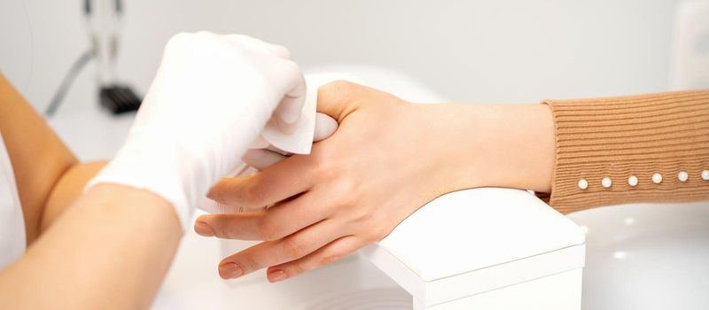 Hands of a manicurist in white protective gloves wipe female nails with a paper napkin in the salon