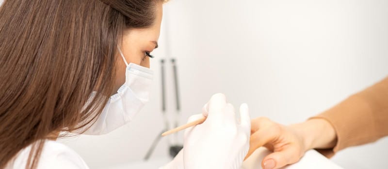 Manicure master in protective face mask and white gloves apply polish to female fingernails