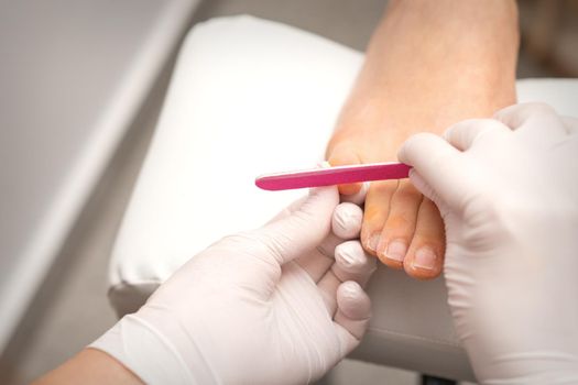 Pedicurist hands in protective rubber gloves filing toenails on feet with a nail file in a beauty salon