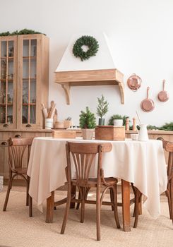 Modern, light, festive, cozy kitchen interior with Christmas and New Year decorations, kitchen table, utensils, copper pans on wall and big Christmas tree with presents, winter holidays concept