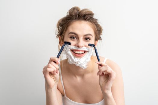 Beautiful young caucasian woman shaving her face by razor on white background. Pretty smiling woman with shaving foam and razor on her face