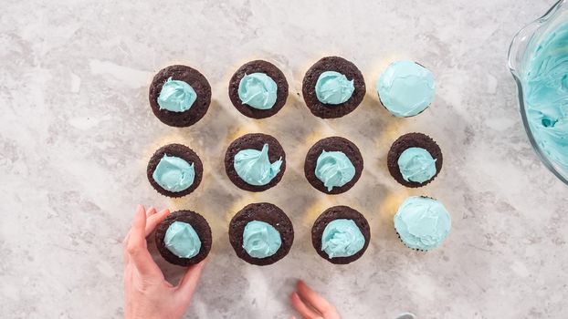 Flat lay. Step by step. Scooping blue buttercream frosting on top of the chocolate cupcakes.