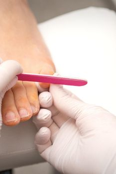 Pedicurist hands in protective rubber gloves filing toenails on feet with a nail file in a beauty salon