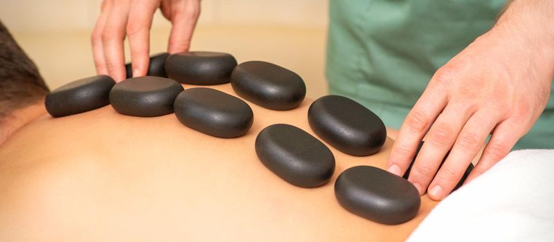 Young caucasian man receiving back massage with black stones by masseur in spa salon. A woman getting a spa treatment