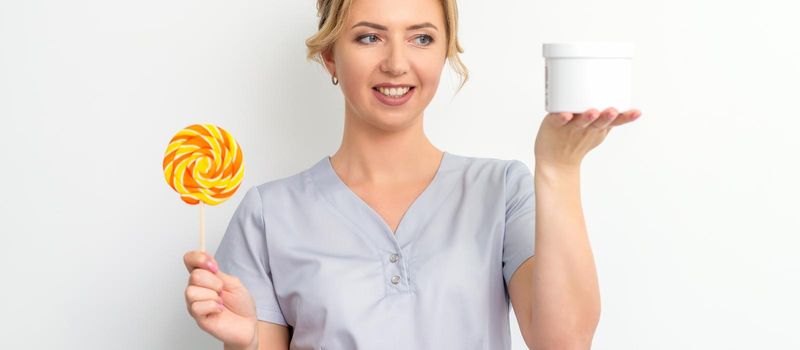 Beautician holding a jar of wax for waxing and candy on a stick smiling on a white background. Natural product for hair removal