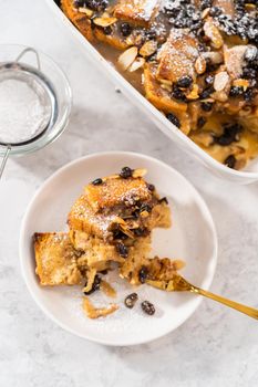 Serving freshly baked bread pudding with raising and almond slivers in a white casserole dish.