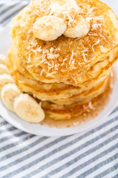 Stack of freshly baked coconut banana pancakes garnished with sliced bananas, toasted coconut, and maple syrup.