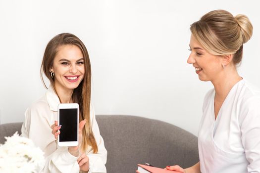 Two smiling female doctors wearing white medical gowns with empty smartphone screen in a medical office, mockup, copy space
