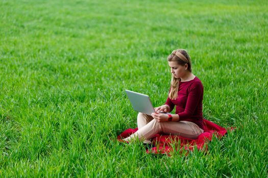 Young successful woman is sitting on green grass with a laptop in her hands. Work on the nature. Student girl working in a secluded place. New business ideas