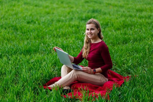 Young successful woman is sitting on green grass with a laptop in her hands. Work on the nature. Student girl working in a secluded place. New business ideas