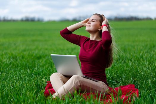 Young successful woman is sitting on green grass with a laptop in her hands. Rest after a good working day. Work on the nature. Student girl working in a secluded place. New business ideas