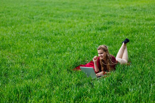 Beautiful young blonde woman is lying on the green grass in the park with a laptop and working. Blue sky with clouds. The girl smiles and enjoys a good day. Work on the nature on a sunny day.