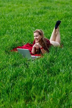 Beautiful young blonde woman is lying on the green grass in the park with a laptop and working. Blue sky with clouds. The girl smiles and enjoys a good day. Work on the nature on a sunny day.