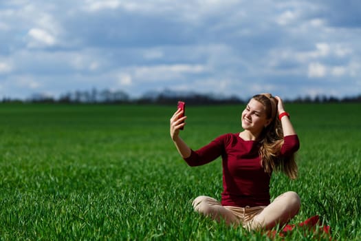 Young beautiful woman student with a phone in her hands sitting on the grass. Girl takes selfies and takes selfie pictures. She smiles and enjoys a warm day. Concept photo on smartphone