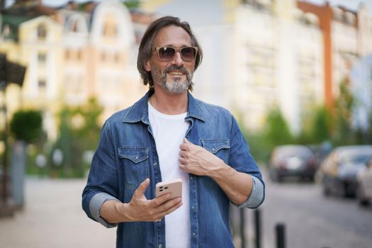 Mature handsome man with smartphone in hand walking on old town streets wearing denim shirt and sunglasses. Handsome middle aged man with grey hair making photos while traveling.