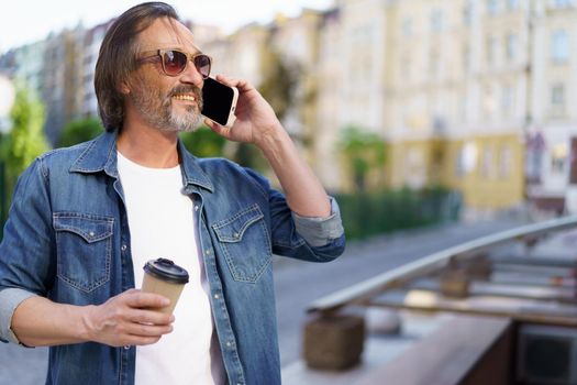 Handsome grey beard middle aged man talking on the phone holding coffee in disposable paper cup standing outdoors in old city background wearing jeans shirt. Freelancer traveling man.