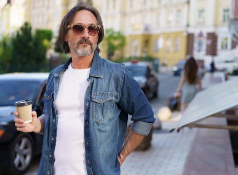 Serious, thoughtful mature man hold paper cup with coffee standing outdoors in old town city wearing jeans shirt. Mature man wearing sunglasses and looking sideways outdoor on a summer day.