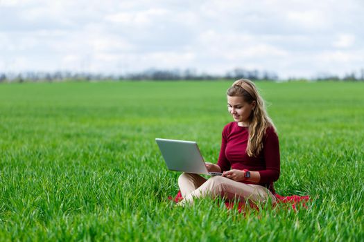 Young successful woman is sitting on green grass with a laptop in her hands. Work on the nature. Student girl working in a secluded place. New business ideas