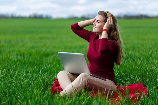 Young successful woman is sitting on green grass with a laptop in her hands. Rest after a good working day. Work on the nature. Student girl working in a secluded place. New business ideas