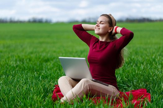 Young successful woman is sitting on green grass with a laptop in her hands. Rest after a good working day. Work on the nature. Student girl working in a secluded place. New business ideas