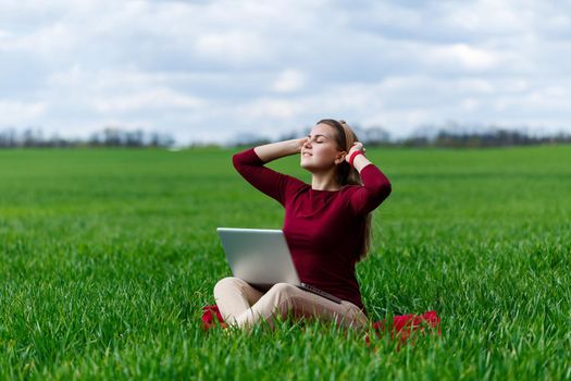 Young successful woman is sitting on green grass with a laptop in her hands. Rest after a good working day. Work on the nature. Student girl working in a secluded place. New business ideas