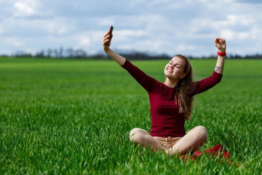 Young beautiful woman student with a phone in her hands sitting on the grass. Girl takes selfies and takes selfie pictures. She smiles and enjoys a warm day. Concept photo on smartphone