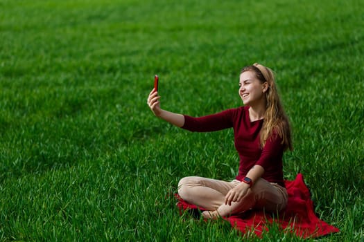 Beautiful woman with a phone in her hands sits on the grass. The girl looks at herself in the camera of the phone and takes selfie photos. She smiles and enjoys a warm day. Concept photo on smartphone