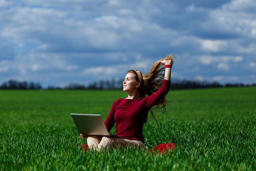 Joyful young girl on the grass with a laptop on her lap. He raised his hands up and laughed. Happiness in the lifestyle of a classical student. Work on the nature. Rest after a good working day.