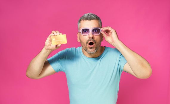Excited of a new offer handsome man hold debit, credit card in hand lowering his blue glasses wearing blue t-shirt isolated on pink background. Man with bank card in hand. Financial, banking concept.
