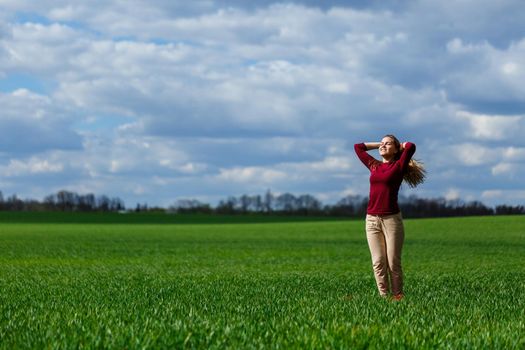 Beautiful young blonde woman stands on green grass in a park. Blue sky with clouds. The girl smiles and enjoys a good day.